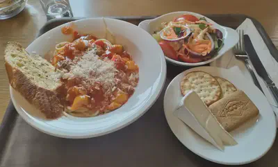 A white plate of spaghetti with red and yellow vegetables, tomato sauce, grated cheese on top, and a slice of garlic bread on the rim. Next to this is a small white casserole dish of salad and a small white plate with brie and crackers.