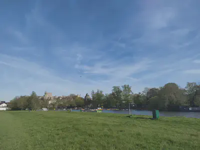 River running across the width of the photo. Grassy foreground and blue sky with an airplane overhead. Windsor Castle in the left background.
