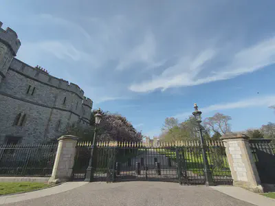 Black gates with lampposts and a high castle wall on the left behind the gates.