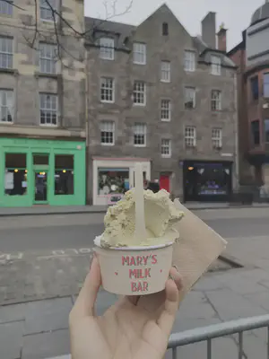 A hand holding two scoops of ice cream in a cup that says Mary’s Milk bar and a napkin behind the cup. The front-facing ice cream is green and has a handle sticking out of it. The blurred background is the street on which Mary’s Milk bar is located.
