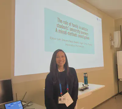 An Asian woman with long brown hair and wearing a navy blue sweater with conference lanyard around her neck stands in front of two desks which are against a wall. The title slide of a presentation is projected onto the wall. Slide title is The role of family in autistic students’ university journeys colon a mixed methods investigation. Slide subtitle is author information which reads Elinor Lim, Steven Kapp, Beatriz Lopez, Emine Gurbuz, University of Portsmouth.