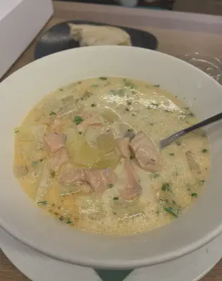 A large white bowl of beige liquid with chunks of pink salmon and yellow potato. Chopped green herbs float on the surface of the soup. In the blurry background is a slice of white bread on a black plate.